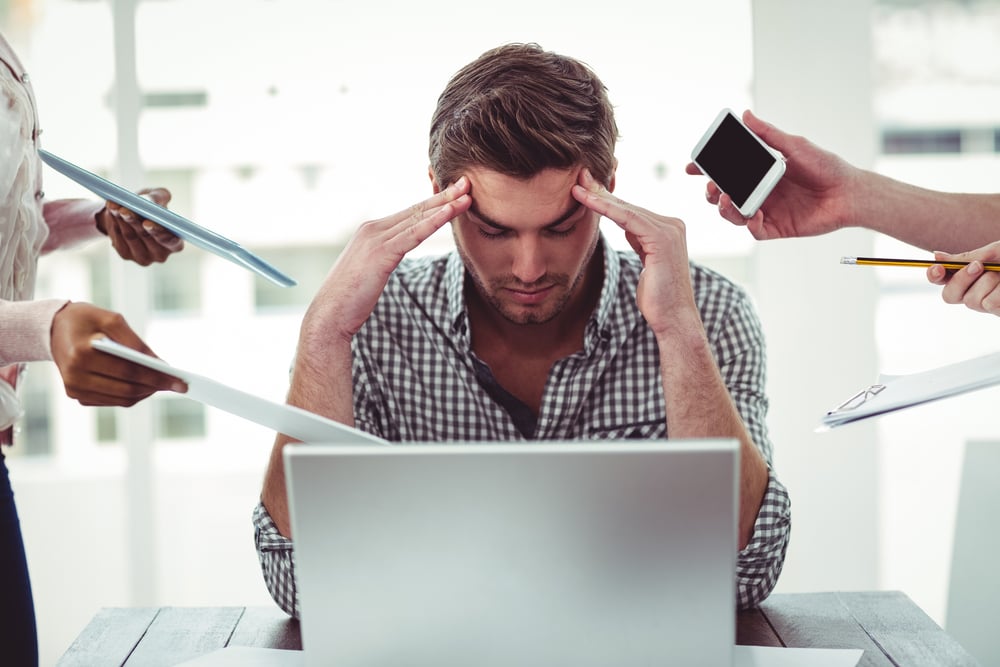 A white man with brown hair and beard wearing a brown chequered shirt sat at a laptop with his head in his hands. Hands with paper, phones, pens, reports are coming at him from both sides.