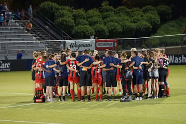 a diverse sports team gathered in a huddle, on the pitch with the coach leading a motivational talk. 